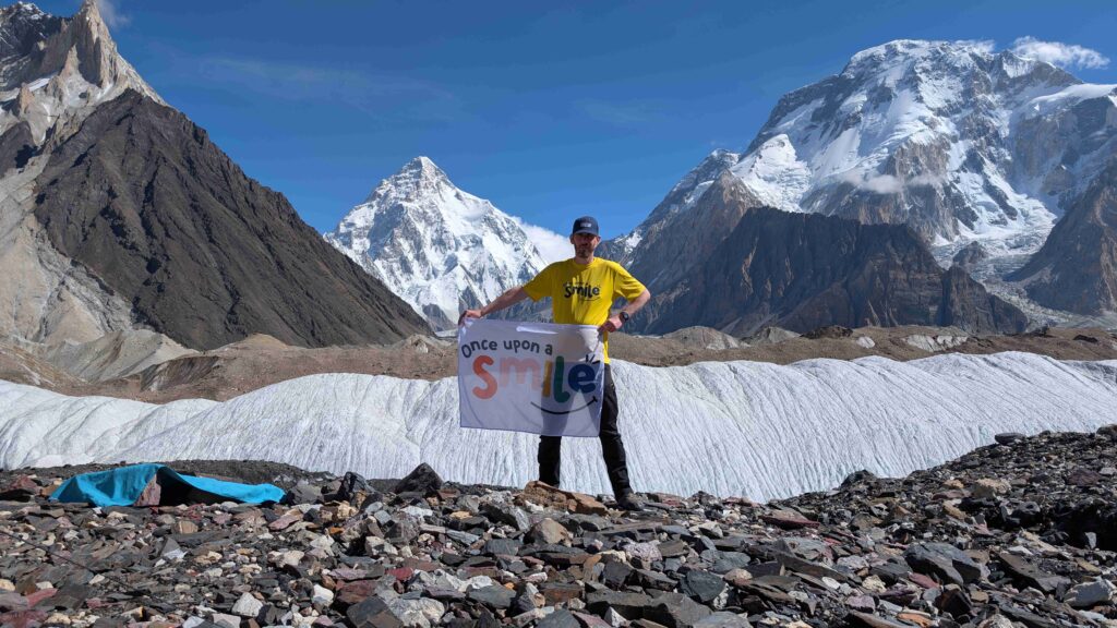 A man stood holding a flag with the Once Upon a Smile logo on with the backdrop of large mountains with snow caps and blue skies